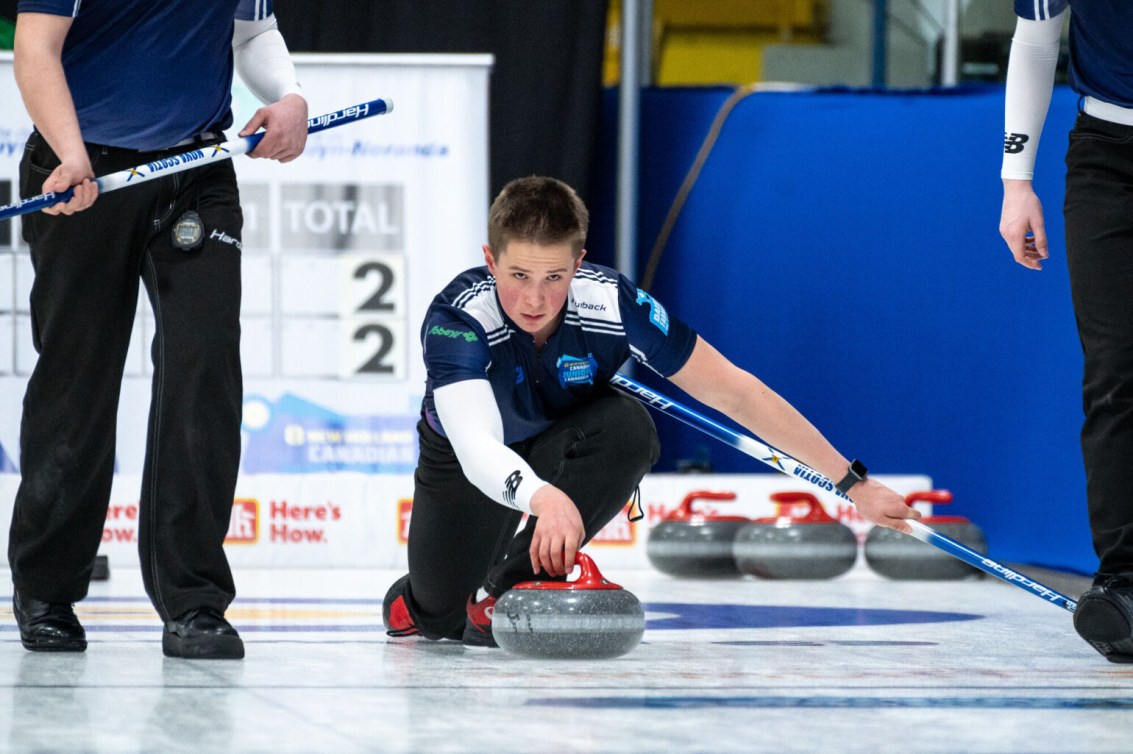 Curler in blue shirt slides to throw a red stone