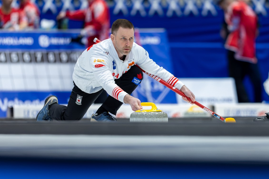 Brad Gushue in a white jacket slides to throw a yellow curling stone