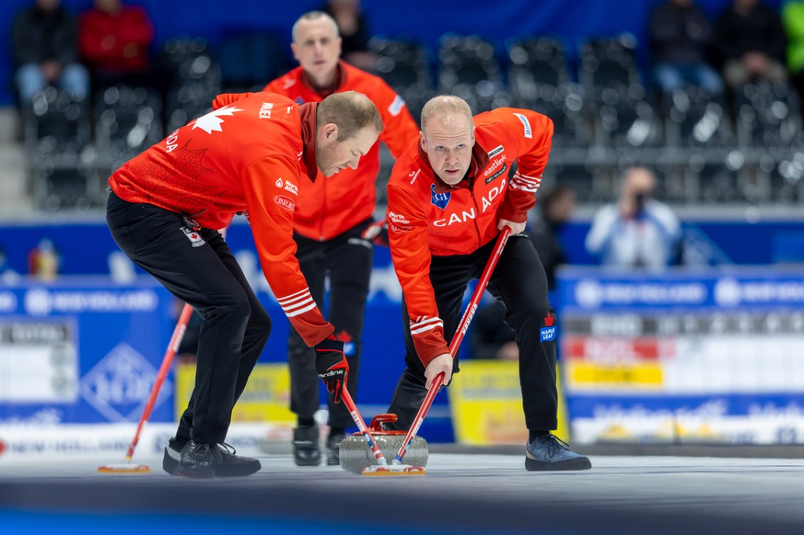 Two curlers in red jackets sweep a stone as their teammate looks on
