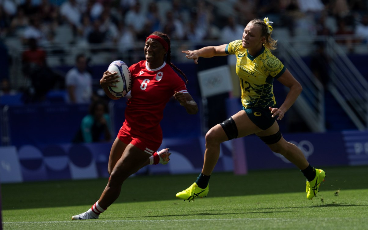 Charity Williams in a red uniform runs with the rugby ball as an Australian player chases her