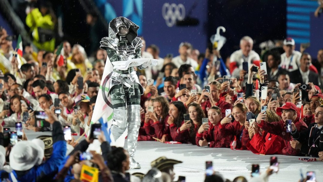 Team Canada athletes stand alongside a stage as a woman in a metal outfit carries the Olympic flag