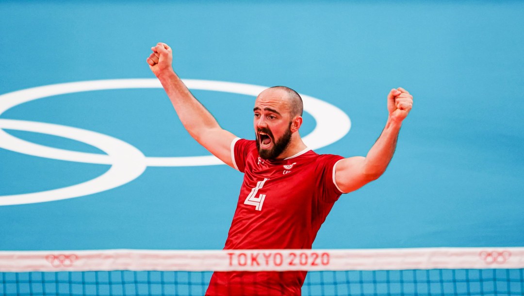 Nicholas Hoag raises both arms in the air in celebration while standing on a blue volleyball court