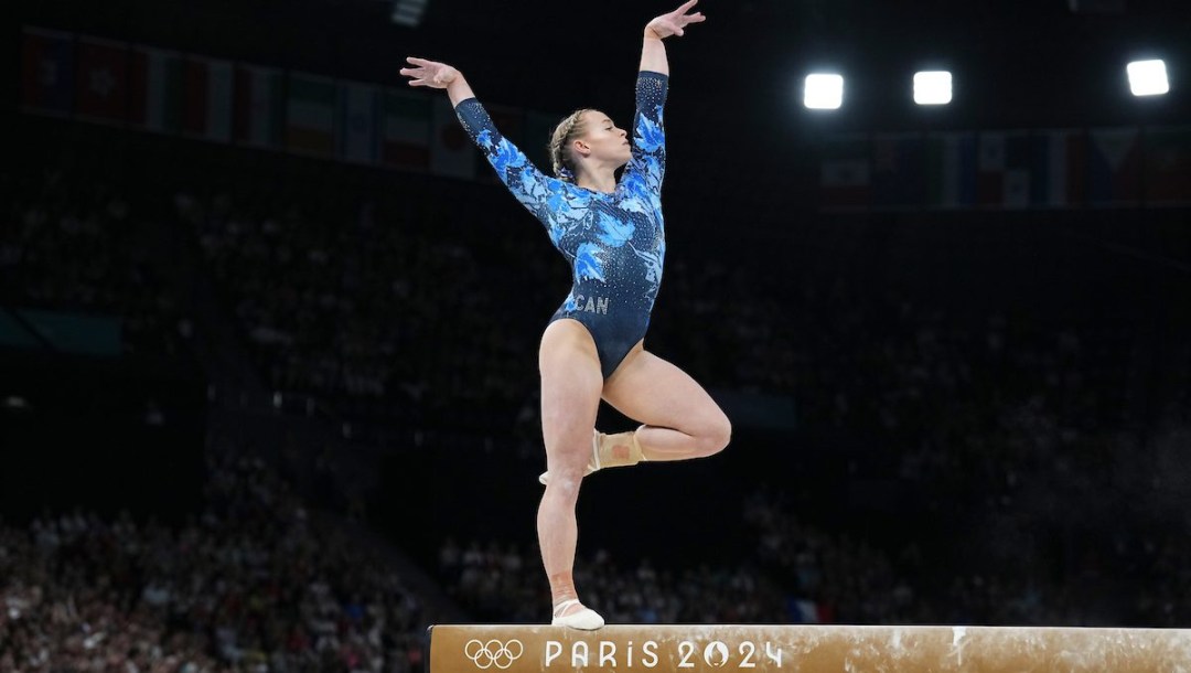 Ellie Black in a blue leotard poses on one leg with her arms in the air on balance beam