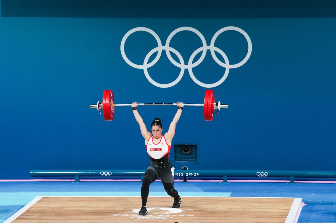 Maude Charron holds a barbell above her head in front of a blue background with the Olympic rings on it