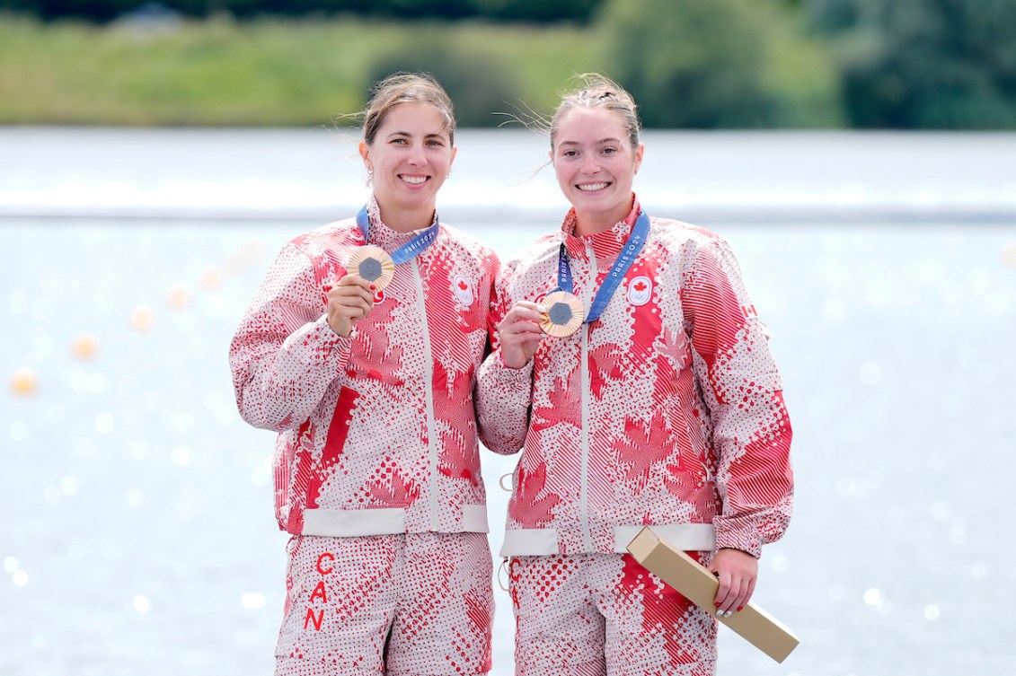 Katie Vincent and Sloan Mackenzie pose while wearing bronze medals with red a white tracksuits