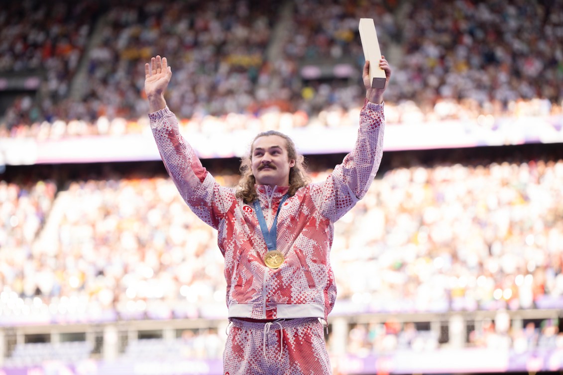 Ethan Katzberg raises his arms after receiving his gold medal in a red and white tracksuit