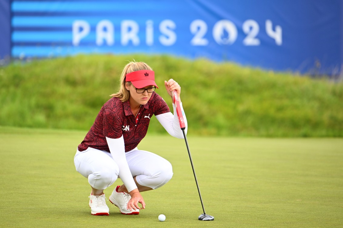 Brooke Henderson squats to line up her putt while wearing a red shirt and white pants