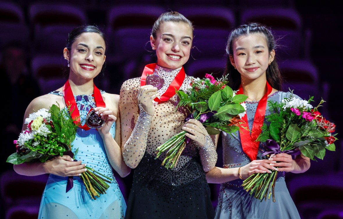 Maddie Schizas in a light blue dress, Kaiya Ruiter in a beige and black dress, and Hetty Shi in a grey dress wear medals on the podium