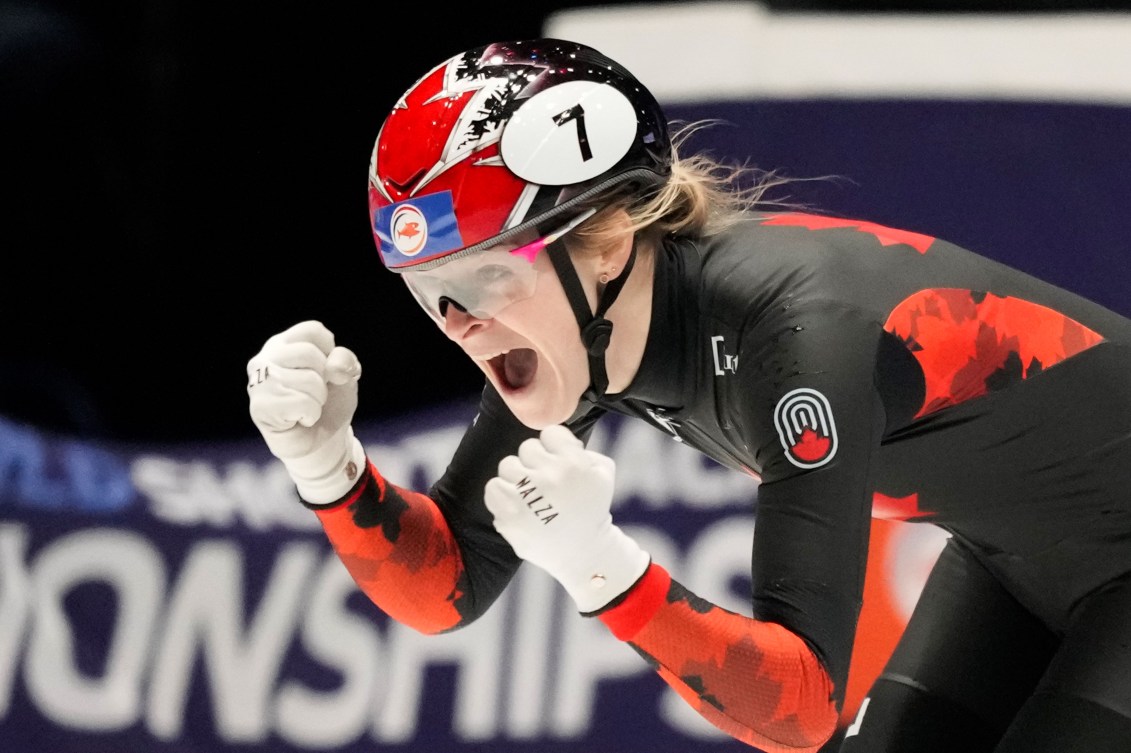 Kim Boutin pumps her fists and yells in victory while wearing a black and red speed skating suit and black and red helmet