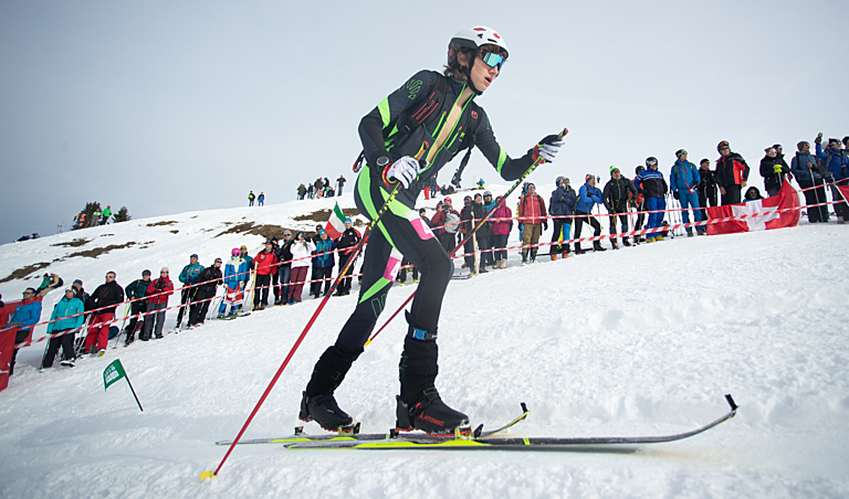 A skier dress in black with green trim on his suit pushes past spectators on a hill 