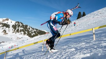 A skier dressed in blue and wearing a helmet walks uphill on snow carrying their skis on their backpack