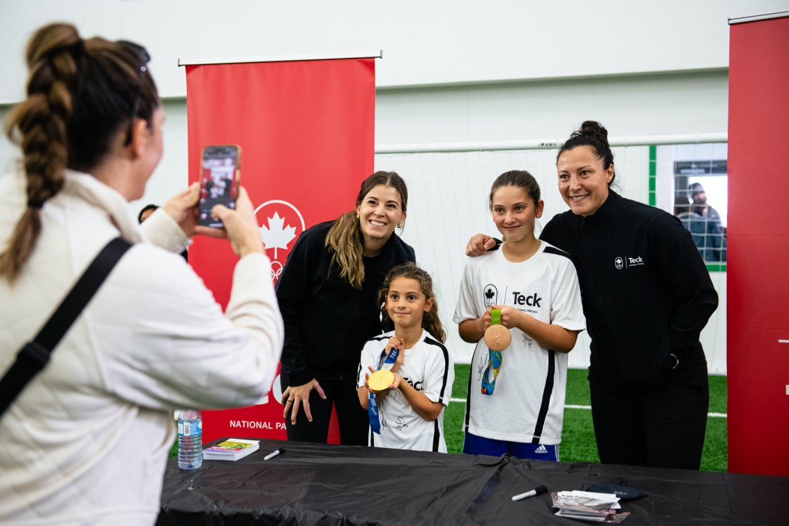 Melissa Tancredi and Steph Labbé pose with youth holding their Olympic medals while a parent takes a photo