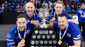 Four male curlers in blue shirts huddle around the Brier trophy