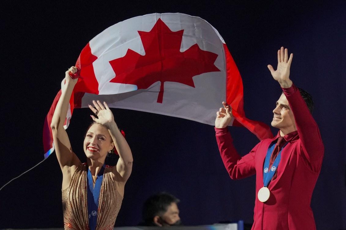 Piper Gilles in a gold dress and Paul Poirier in a red suit carry a Canadian flag between them