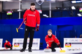 two people on a sheet of ice, man stands wearing red jacket holding curling broom, woman wearing red jacket slides on ice pushing curling rock ahead of her