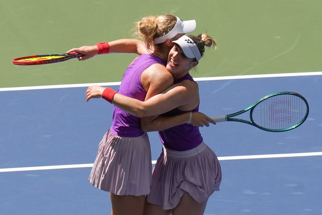 Wearing purple tank tops and pink skirts two tennis players hug each other on court 