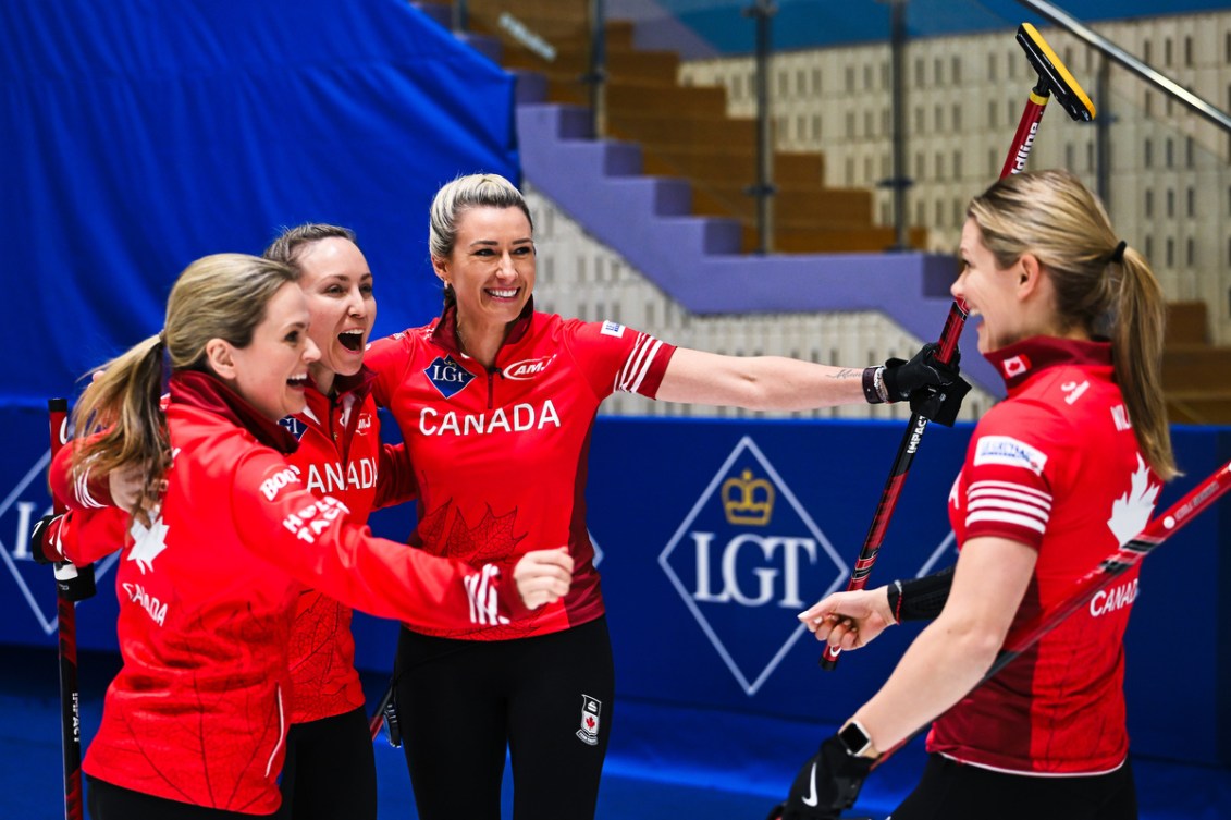 Three women curlers in red jackets prepare to welcome their teammate into a group hug