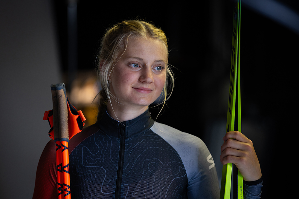 Sonjia Schmidt wearing a black and grey skinsuit holding a pair of neon green skis and neon orange ski poles.  