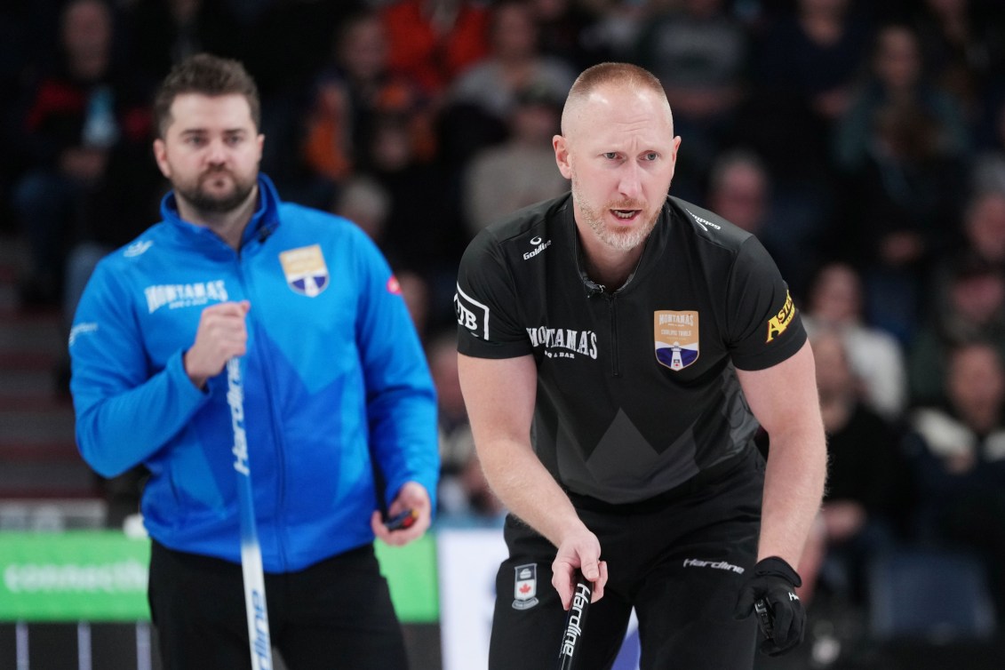 A male curler in blue jacket stands behind a male curler in black shirt 