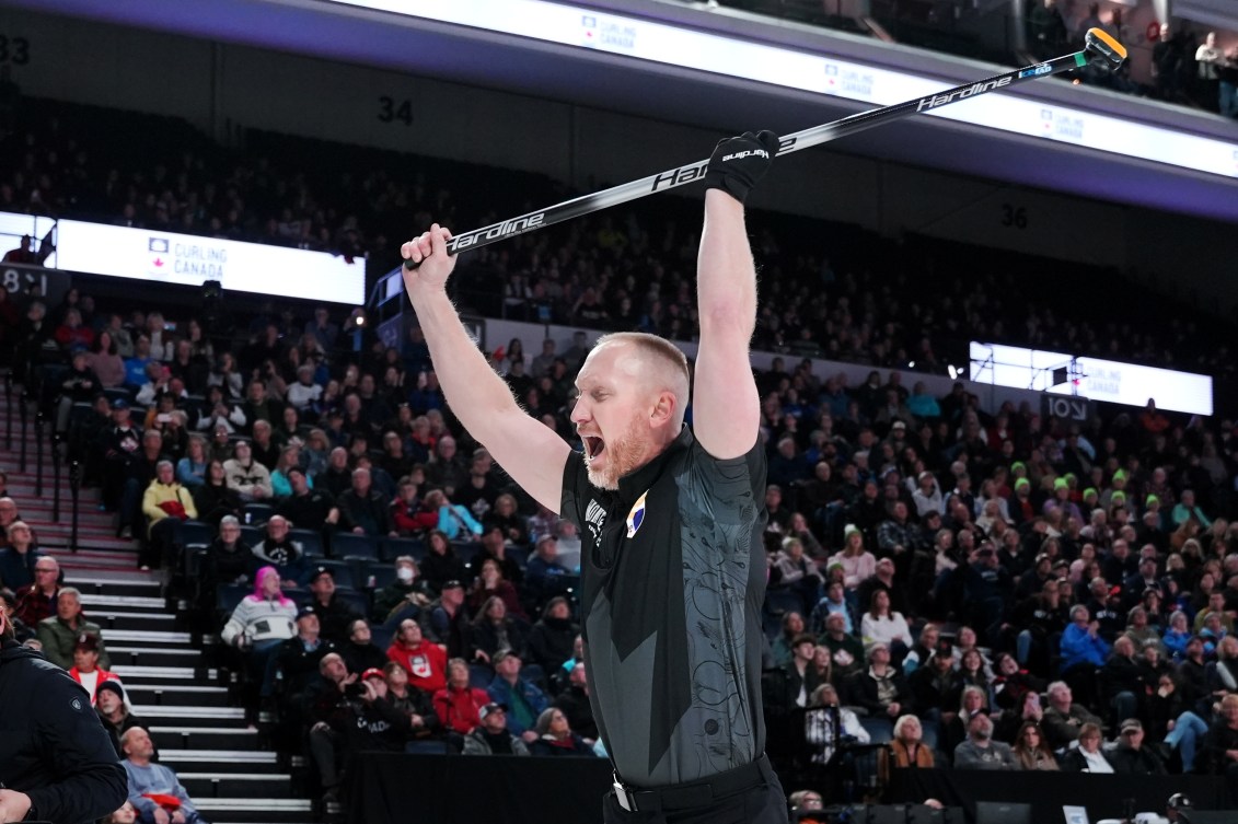 Brad Jacobs in a black shirt raises his broom above his head while screaming in happiness