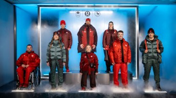Eight athletes stand in front of blue lit up background in various items of the lululemon clothing kit for Milano Cortina 2026 in shades of reds and greens