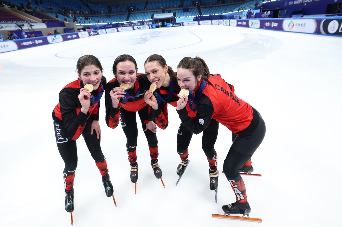 Four female short track speed skaters bite gold medals standing on the ice 