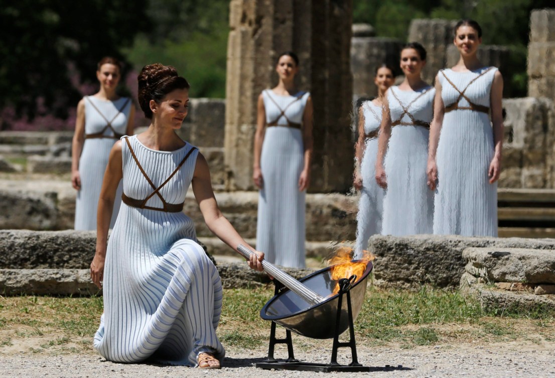 Une femme en robe blanche allume la flamme dans un décor de la Grèce antique